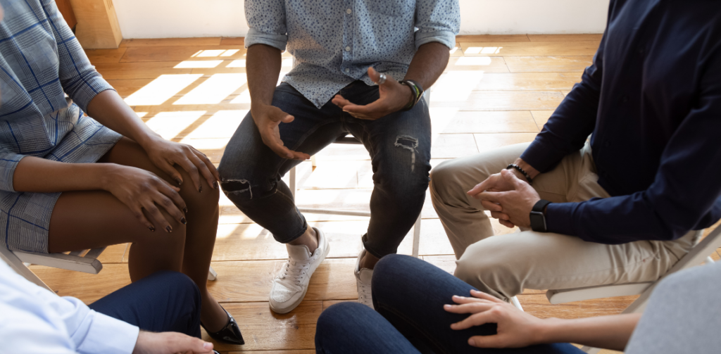 Photo shows a group of people sitting together in a circle from the chest down. They are in discussion with one another.