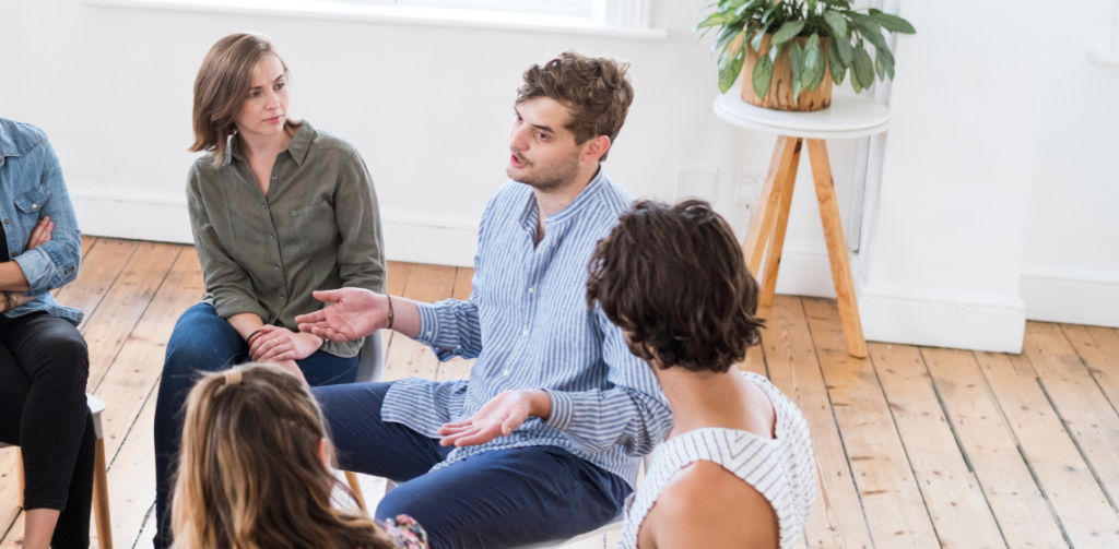 Three people are seated in a larger group of people in a circle, one person is speaking while two others listen intently